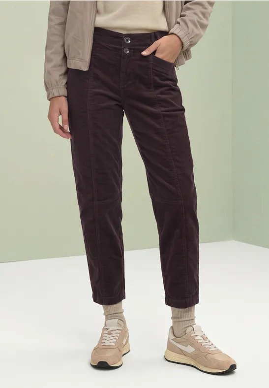 Eine Person trägt dunkle Cordhose und beige Sneaker vor neutralem Hintergrund.