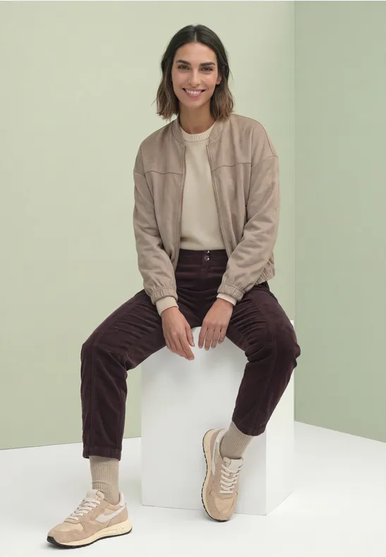 Frau in beige Jacke, weißem Shirt, braunen Cordhosen und beigen Sneakern sitzt auf weißem Würfel vor grünem Hintergrund.
