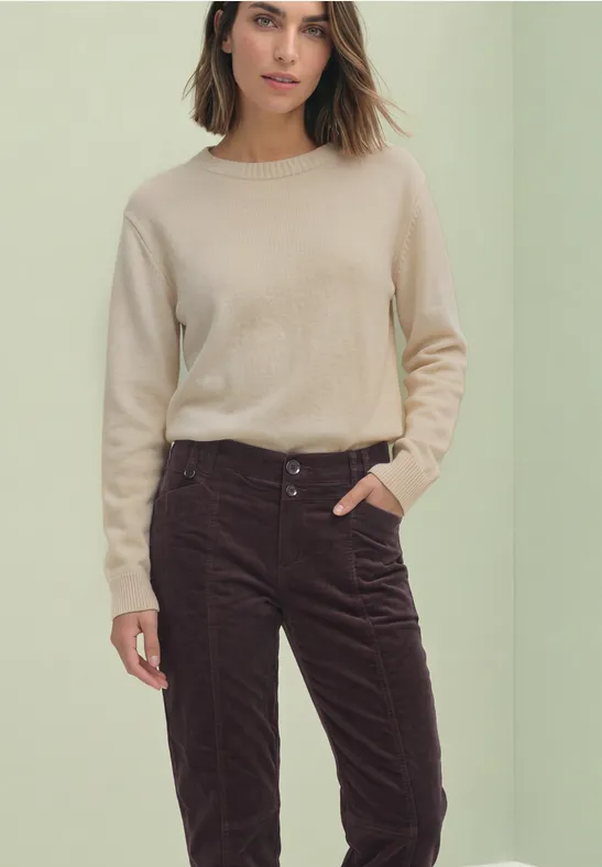 Eine Frau trägt einen beigen Pullover und dunkelbraune Cordhose, Hand in Tasche.