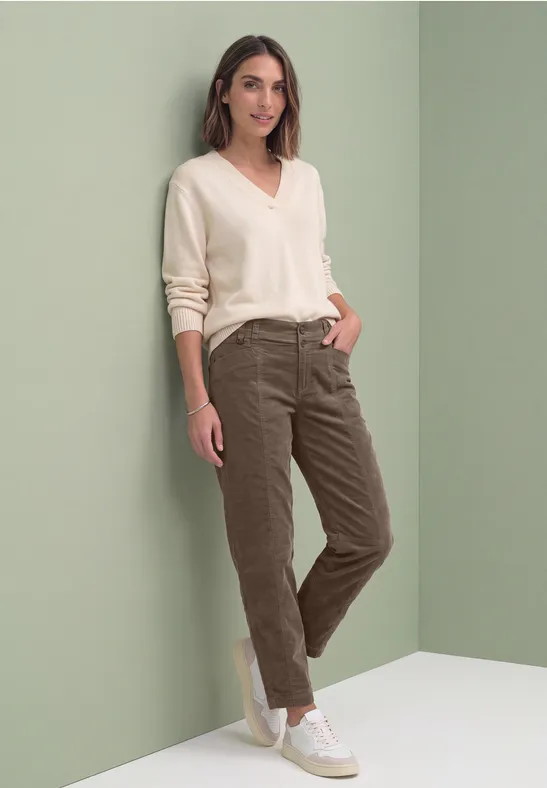 Vrouw leunt ontspannen tegen een lichtgroene muur, draagt beige trui, kaki broek en witte sneakers, glimlacht.