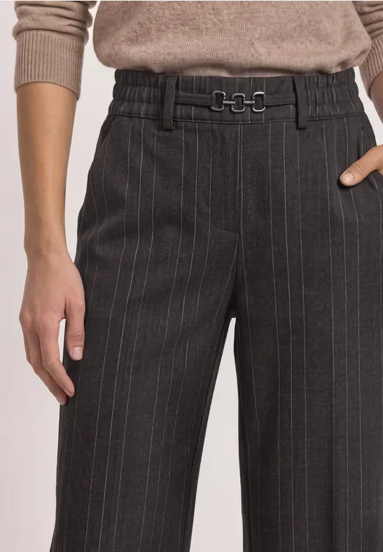 Dunkelgraue Nadelstreifenhose mit weitem Bein und feinem Gürtel, kombiniert mit einem beigefarbenen Pullover.
