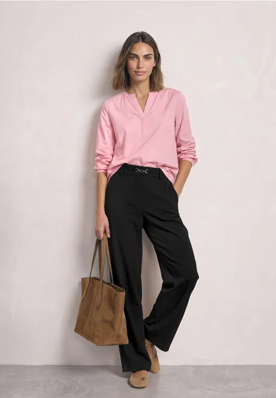 Vrouw in roze blouse en zwarte wijde broek met een bruine tas, staand voor een lichtgrijze achtergrond.