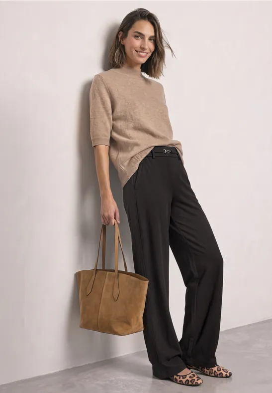 Vrouw in beige trui, zwarte wijde broek, suède tas en luipaardprint ballerina’s leunt tegen een muur.