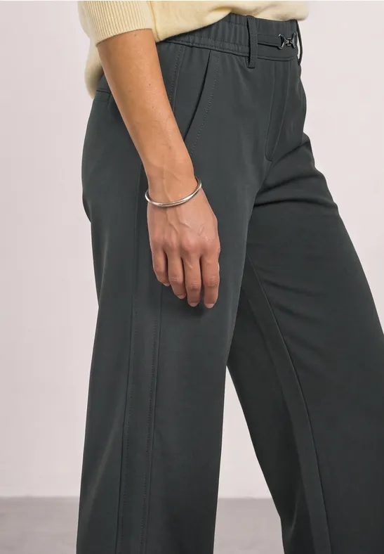 Persoon draagt donkergrijze broek en crème top, hand met zilveren armband zichtbaar.