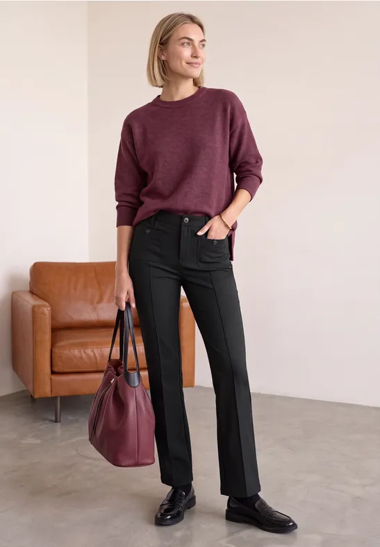 Frau in burgunderrotem Pullover, schwarzer Hose und Schuhen, mit passender Tasche, steht neben braunem Sessel.