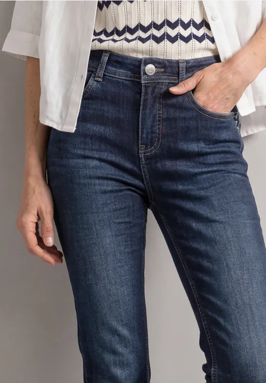 Nahaufnahme einer Person in dunkelblauen Jeans mit Hand in der Tasche, weißem Hemd und gemustertem Oberteil.