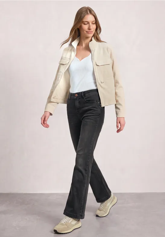 Frau in cremefarbener Jacke, weißem Top, schwarzen Jeans und Sneakers vor neutralem Hintergrund.