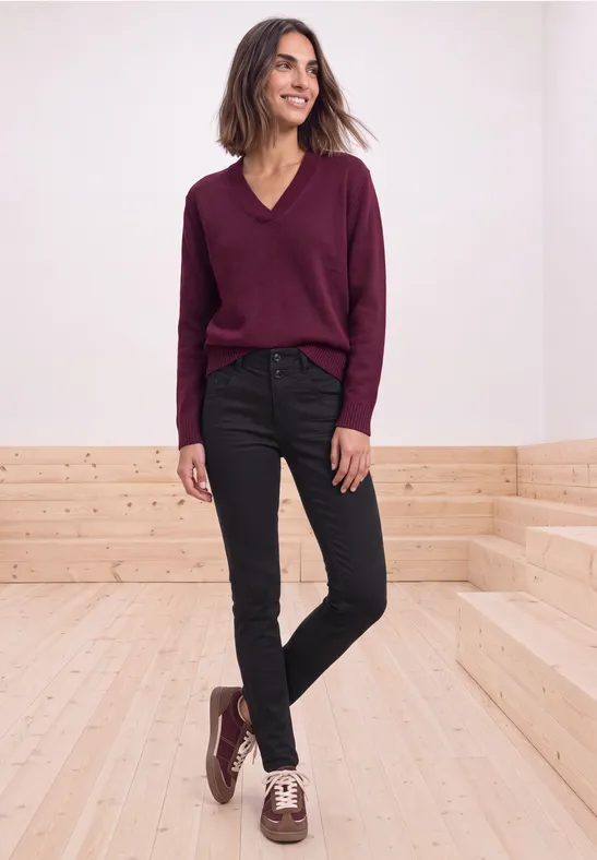 Frau in weinrotem V-Pullover, schwarzen Jeans und zweifarbigen Schuhen steht lächelnd auf Holzstufen vor weißer Wand.