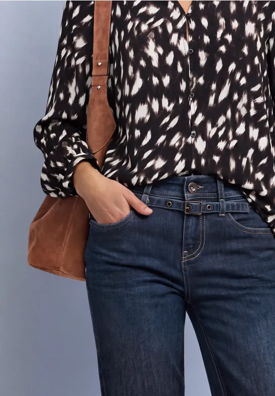 Frau mit schwarz-weiß gemusterter Bluse, blauen Jeans und Umhängetasche, eine Hand in der Tasche.