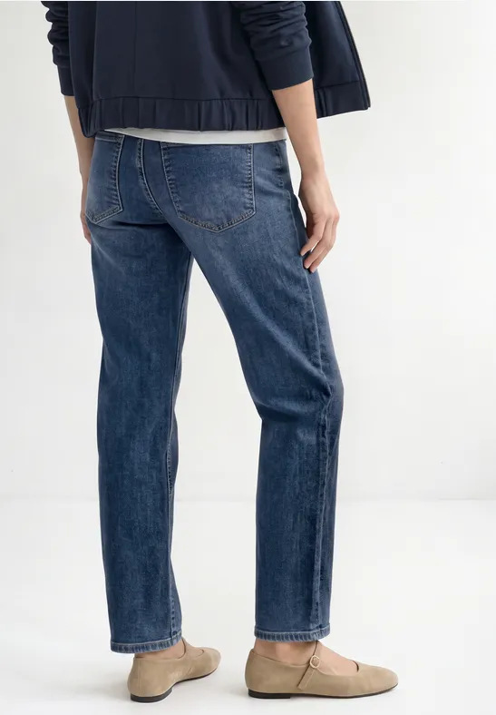Rückansicht einer Person in blauen Jeans, dunklem Oberteil und beigen Halbschuhen.