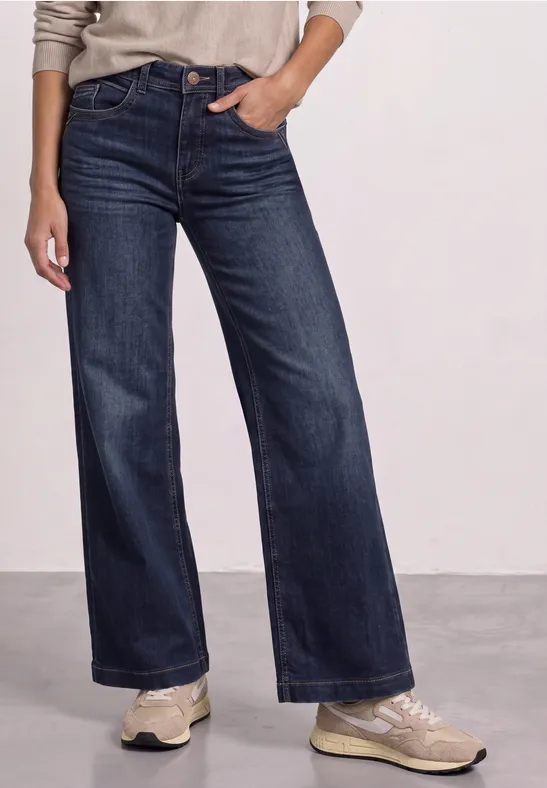 Person trägt weite dunkelblaue Jeans und helle Sneaker.
