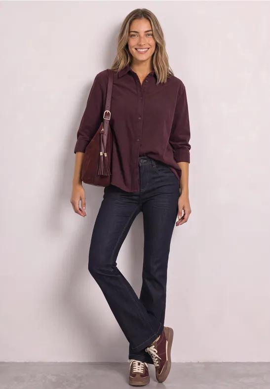 Eine Frau vor hellem Hintergrund trägt weinrotes Hemd, schwarze Jeans, passende Sneaker und eine Umhängetasche.