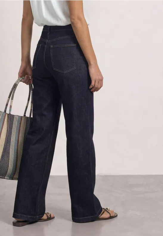 Frau von hinten in hochgeschnittener, weiter, dunkelblauer Jeans, trägt Sandalen und eine gestreifte Tasche.