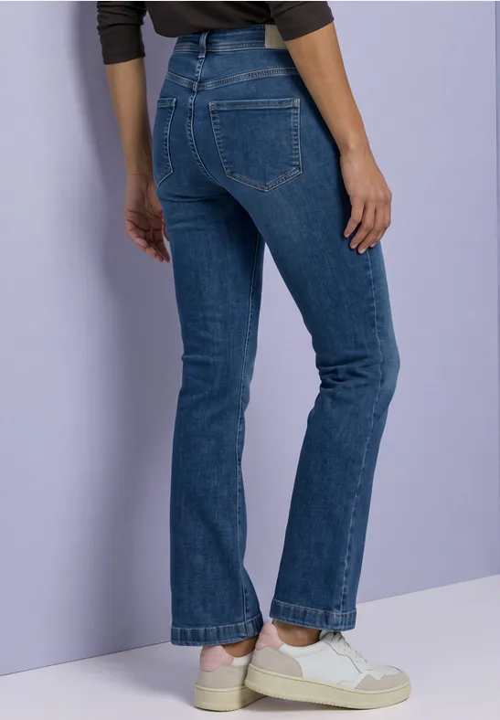 Eine Person steht seitlich, trägt blaue Jeans und weiße Sneaker vor einem hellvioletten Hintergrund.