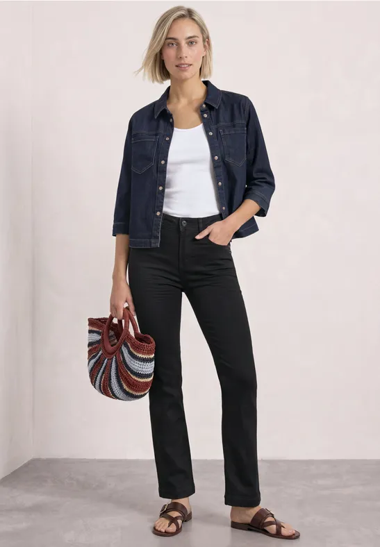 Frau in Jeansjacke, weißem Top, schwarzen Hosen und Sandalen hält bunte Tasche.