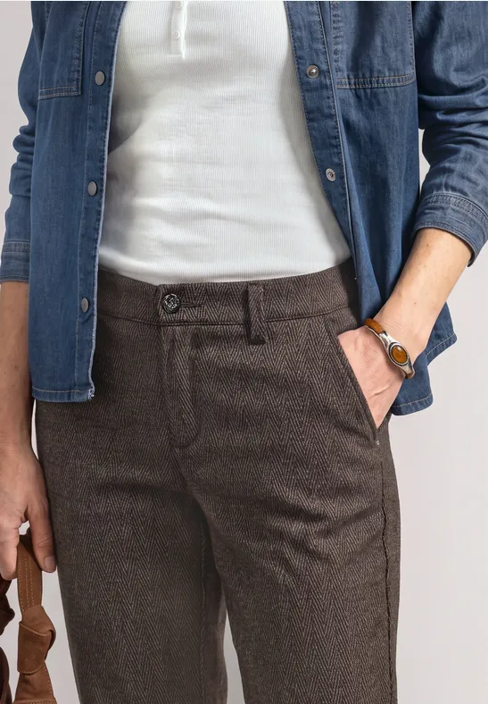 Frau trägt weiße Bluse, offene Jeansjacke und braune Hose, Hand in Tasche mit Armbanduhr.