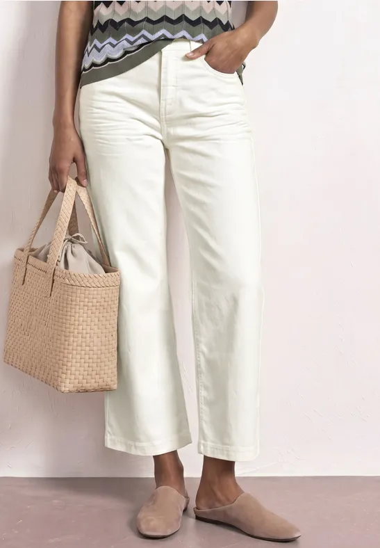 Eine Frau trägt cremefarbene, weite Cropped-Hosen, flache Pantoletten und hält eine gewebte Tasche.