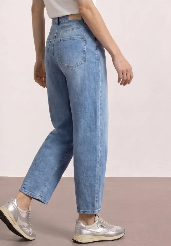 Seitenansicht einer Person in hellblauen, locker sitzenden Jeans und silbernen Sneakern vor neutralem Hintergrund.