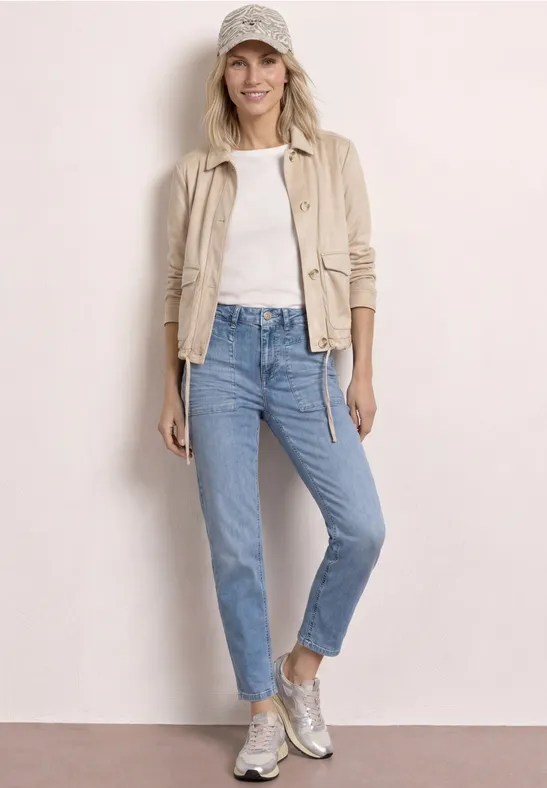 Frau trägt beige Jacke, weißes Shirt, hellblaue Jeans, silberne Sneaker und Basecap.