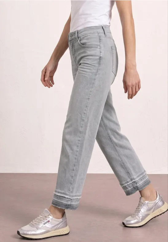 Zijaanzicht van een persoon in lichtgrijze, rechte jeans met omgeslagen pijpen en zilverkleurige sneakers.