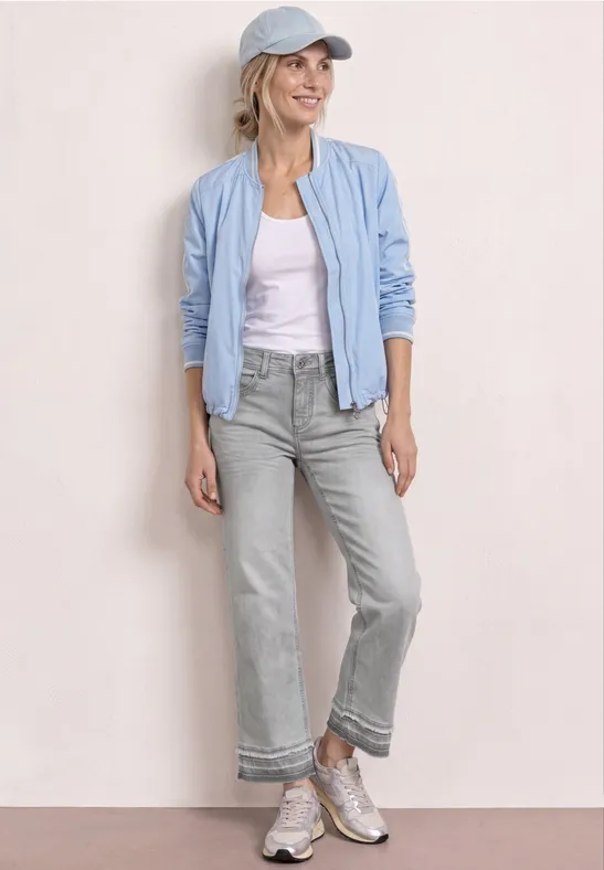 Persoon met lichtblauw bomberjack, wit shirt, grijze cropped jeans, sneakers en pet voor een neutrale achtergrond.