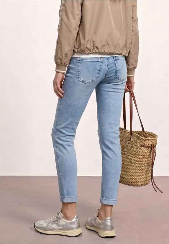Rückenansicht einer Person in hellblauen Jeans, silbernen Sneakers, beiger Jacke und geflochtene Korbtasche.