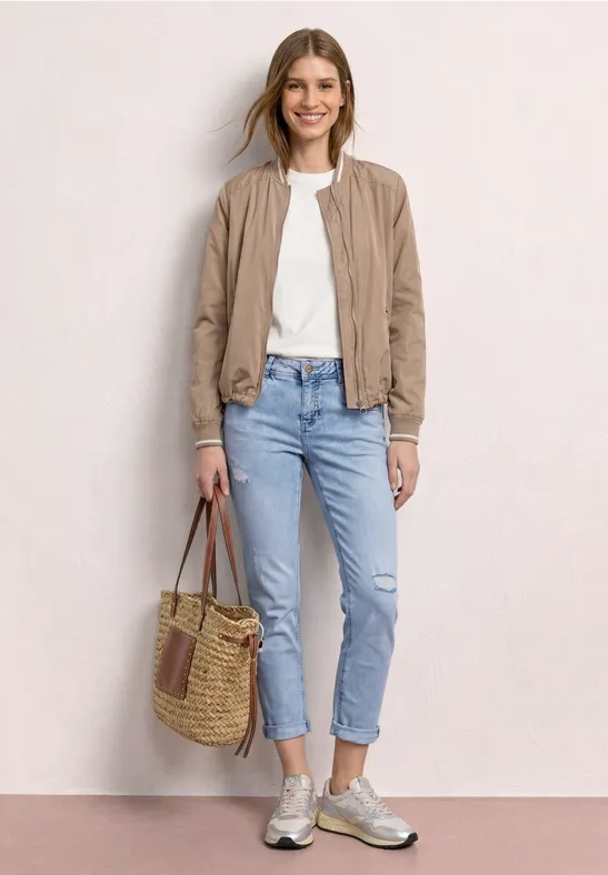 Dame in beiger Jacke, weißem Shirt, blauen Jeans und Sneakers mit geflochtener Tasche vor hellem Hintergrund.