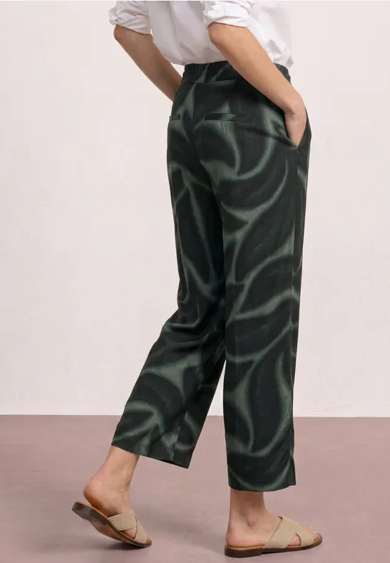 Person trägt weite, dunkelgrün gemusterte Hose mit abstrakten Wirbeln und flache Sandalen.