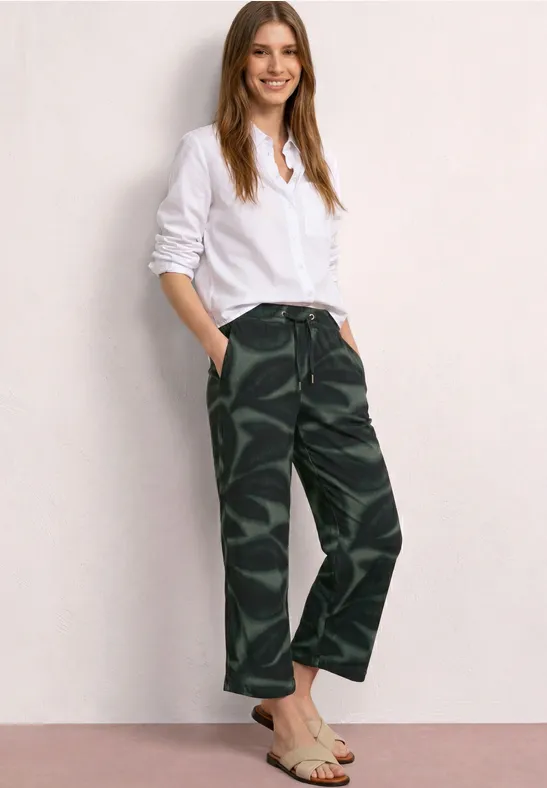 Eine Frau in weißem Hemd und Camouflage-Hose lehnt vor einer hellen Wand.