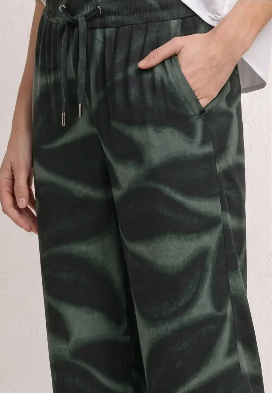 Eine Nahaufnahme einer Person in grün-schwarzen, gemusterten Freizeithosen mit Tunnelzug und Hand in der Tasche.