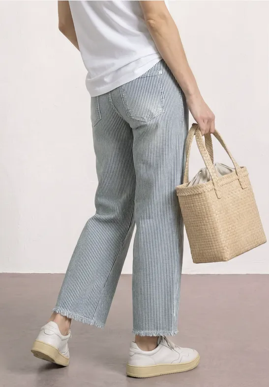 Rückansicht einer Person in weitem Jeans, weißem Shirt und Sneakern, die einen geflochtenen Korb hält.