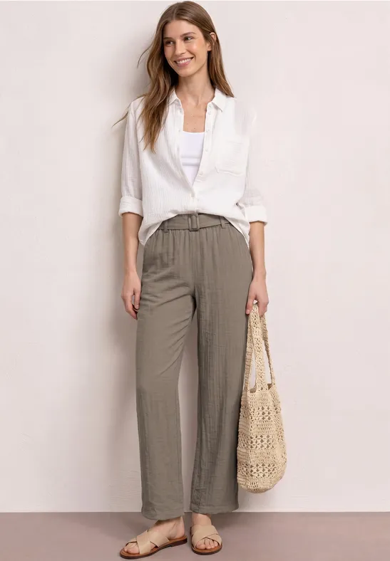 Frau in weißer Bluse, weiten taupefarbenen Hosen und Sandalen, hält einen geflochtenen Korbbeutel.