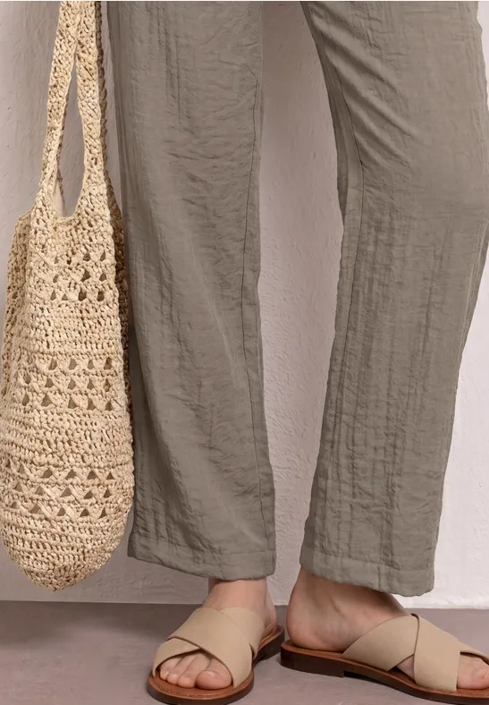 Beine in weiten, cremefarbenen Leinenhosen, beige Riemchensandalen und gehäkelte Einkaufstasche.