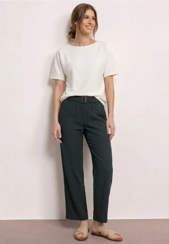 Frau in weißem T-Shirt, dunklen Hose mit Gürtel und Sandalen vor hellem Hintergrund.