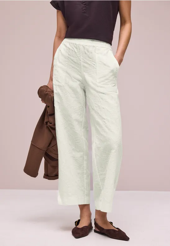 Vrouw in lichte, wijde broek, donker topje ingestopt, met een bruin jasje om de arm en bruine loafers.