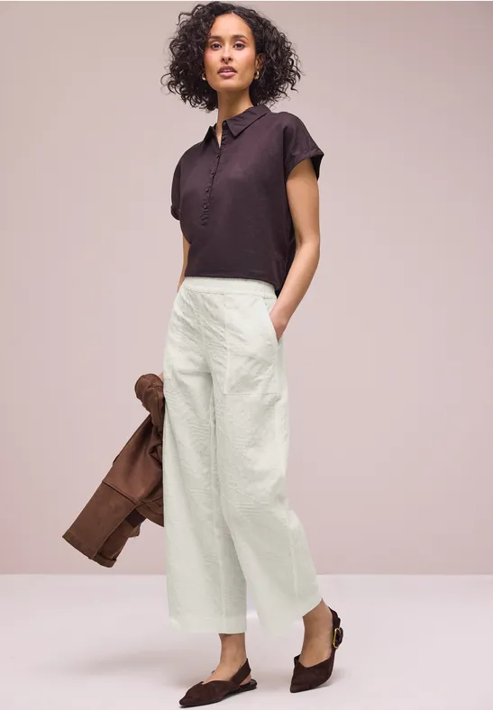 Vrouw in witte, wijdvallende broek en donker poloshirt, met een bruine jas en bruine loafers.