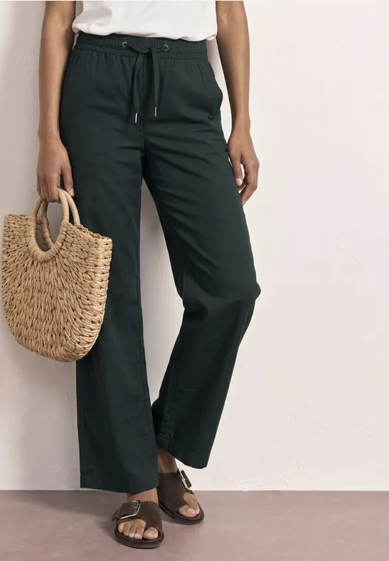 Frau trägt weite, dunkle Leinenhose mit Tunnelzug, flache Sandalen und hält eine geflochtene Strohtasche.