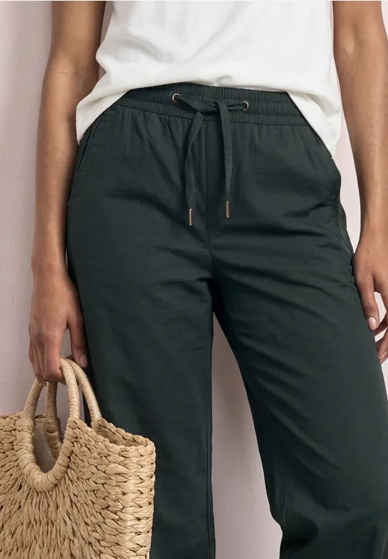 Dunkelgrüne Leinenhose mit Tunnelzug, seitlichen Taschen und Strohtasche in der Hand.