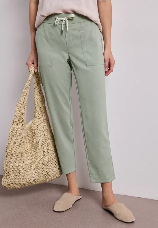 Persoon draagt lichtgroene, rechte broek, beige top, rieten tas en comfortabele instappers.