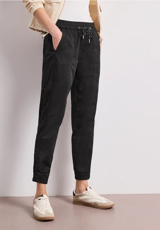 Persoon draagt zwarte, losse jogger met trekkoord en witte sneakers voor een lichte, eenvoudige achtergrond.