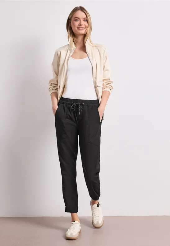 Persoon met een beige hoodie, wit topje, zwarte joggingbroek en witte sneakers voor een lichte achtergrond.