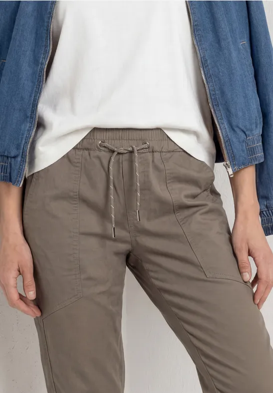 Eine Person trägt graubraune Kordelzug-Hose mit Eingrifftaschen, weißes T-Shirt und Jeansjacke.