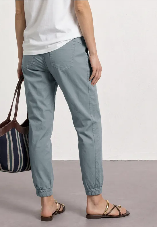 Persoon van achteren in grijze broek met elastische taille, wit shirt, gestreepte tas en platte sandalen.