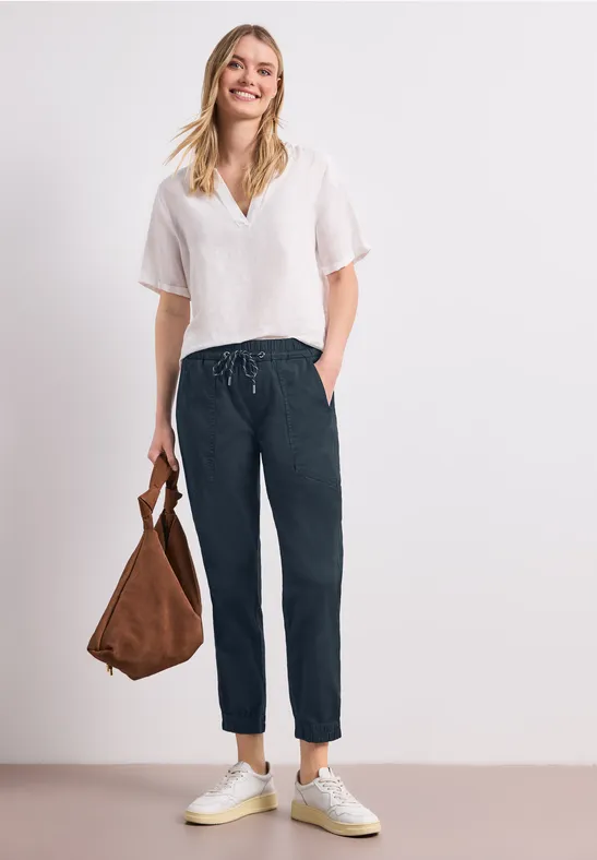 Vrouw in wit linnen topje, donkerblauwe broek, witte sneakers en bruine tas voor een lichte achtergrond.