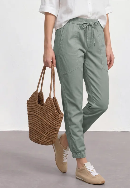 Vrouw in groene joggerbroek met elastische boorden, wit shirt, gevlochten tas en beige sneakers.