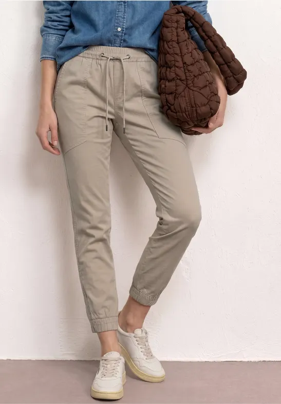 Eine Person trägt bequeme beige Jogginghose, Jeansbluse und weiße Sneakers, hält eine gesteppte braune Tasche.