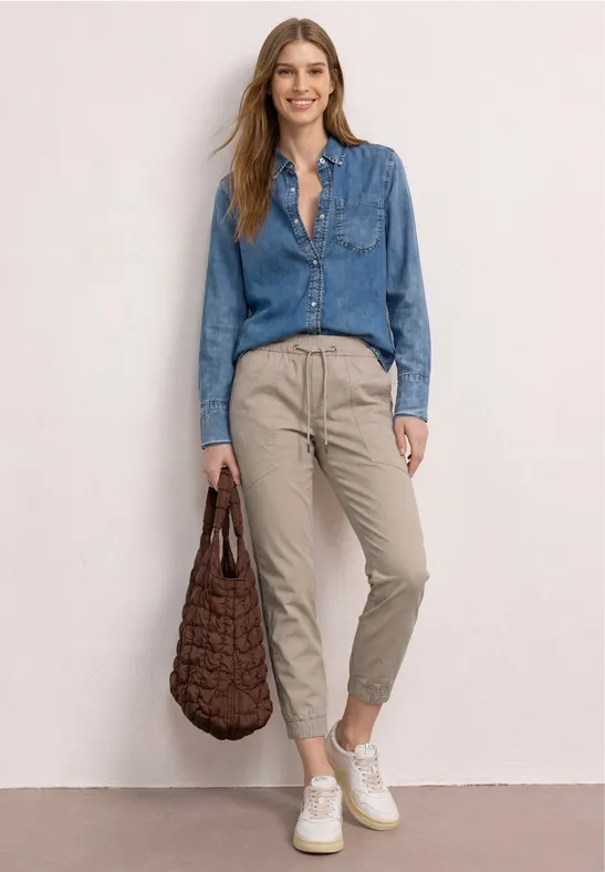Frau trägt Jeanshemd, beige Hose und weiße Sneaker, hält eine geflochtene Tasche.
