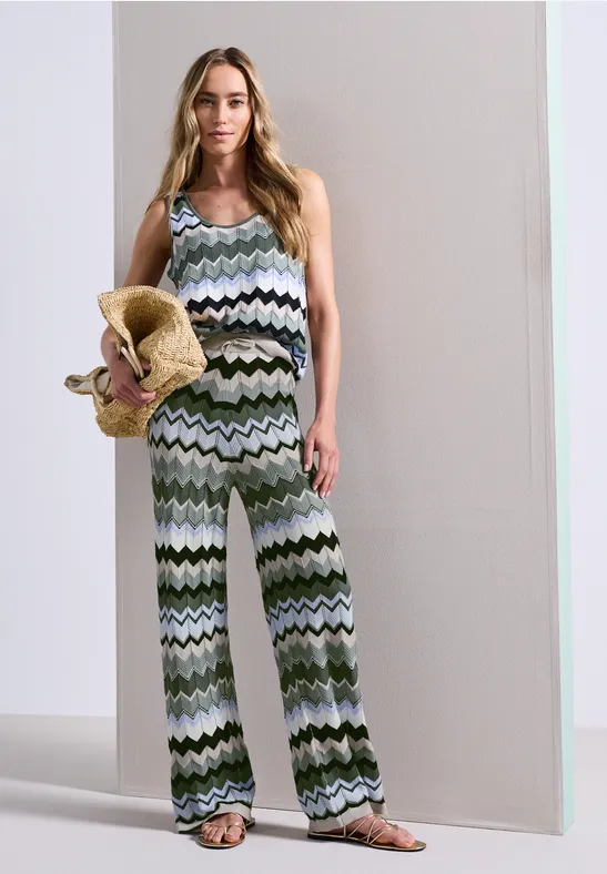 Vrouw in een mouwloze, groen-witte zigzag jumpsuit met wijde pijpen, houdend een strohoed vast.