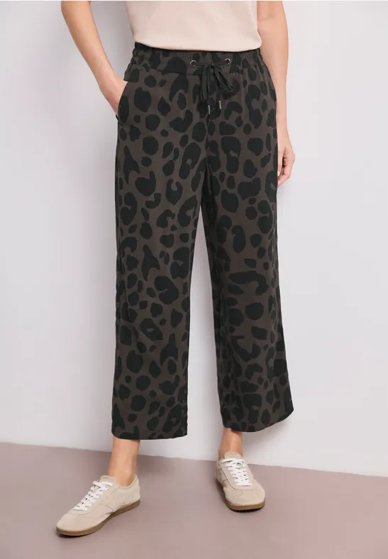 Zwarte, wijdvallende broek met luipaardprint, elastische taille en zakken, gecombineerd met lichte sneakers.