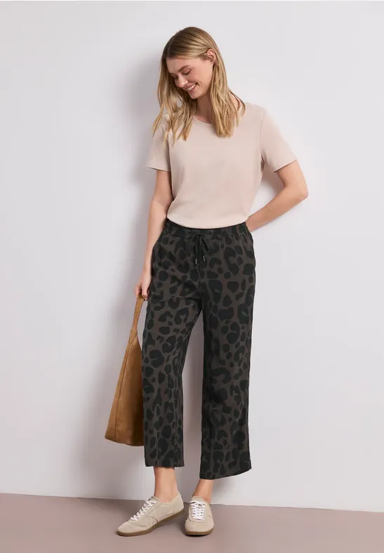 Vrouw in licht t-shirt, wijde broek met dessin, witte sneakers en een eenvoudige tas.